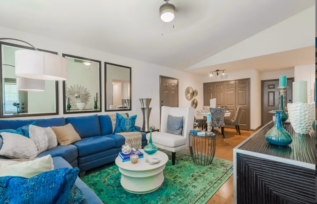 A bright and modern living room with a blue sectional sofa adorned with various pillows, a white armchair, and a round white coffee table on a green patterned rug. The room features three large mirrors on the wall, a ceiling fan, and a dining area with a table and chairs in the background. Decorative vases and candles are placed on a black sideboard and tables.
