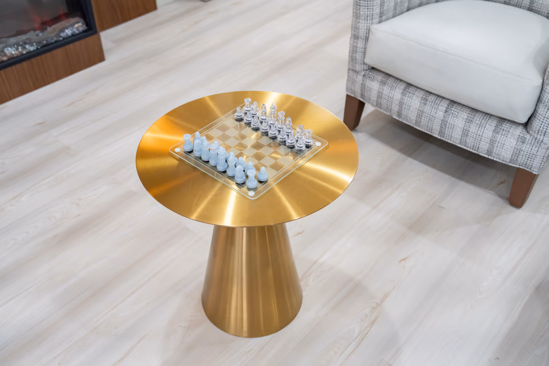 A modern living room area with a small round gold table holding a glass chess set. Next to the table is a gray and white upholstered armchair with a white cushion. The floor is light wood, and a fireplace is partially visible in the background.
