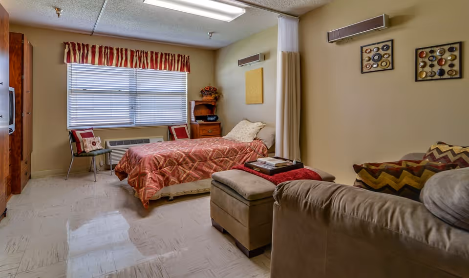 A cozy bedroom in a senior living facility with a single bed covered in a patterned red and beige bedspread. The room has a large window with blinds and a red and beige valance. There are two chairs with decorative pillows near the window, a wooden nightstand with a radio and a flower arrangement, and a beige couch with patterned pillows and an ottoman in the foreground. The walls are decorated with two framed art pieces and a yellow square panel. A ceiling light illuminates the room.