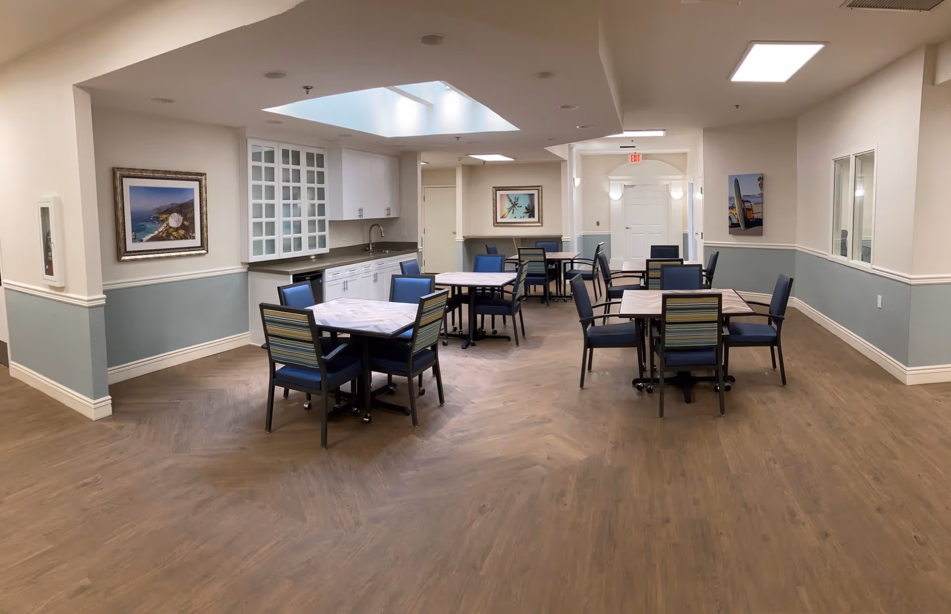 A bright and clean dining room in a senior living facility with several square tables and blue cushioned chairs. The room features wood flooring, white and light blue walls, framed artwork, and a skylight above a kitchenette area with white cabinets and a sink.