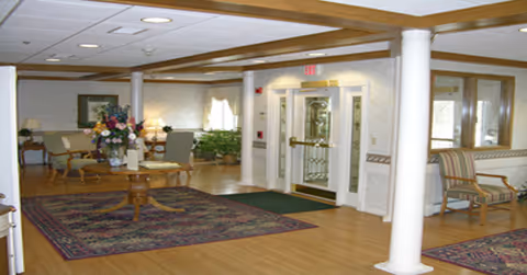 A spacious assisted living facility common area with wooden flooring, two large area rugs, a round wooden table with a floral arrangement in the center, several upholstered chairs, and a glass double door entrance. The room has white walls with wooden trim, ceiling lights, and decorative plants.