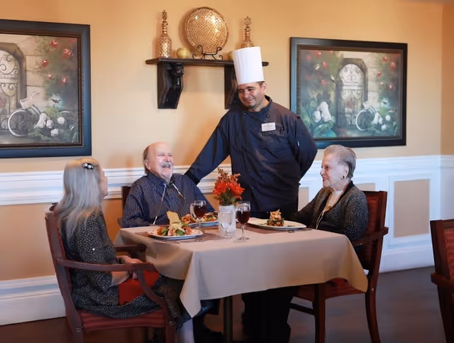 Three elderly people sitting at a dining table with plates of food and drinks, smiling and interacting with a chef standing beside them in a warmly decorated dining room with framed artwork and decorative items on the wall.