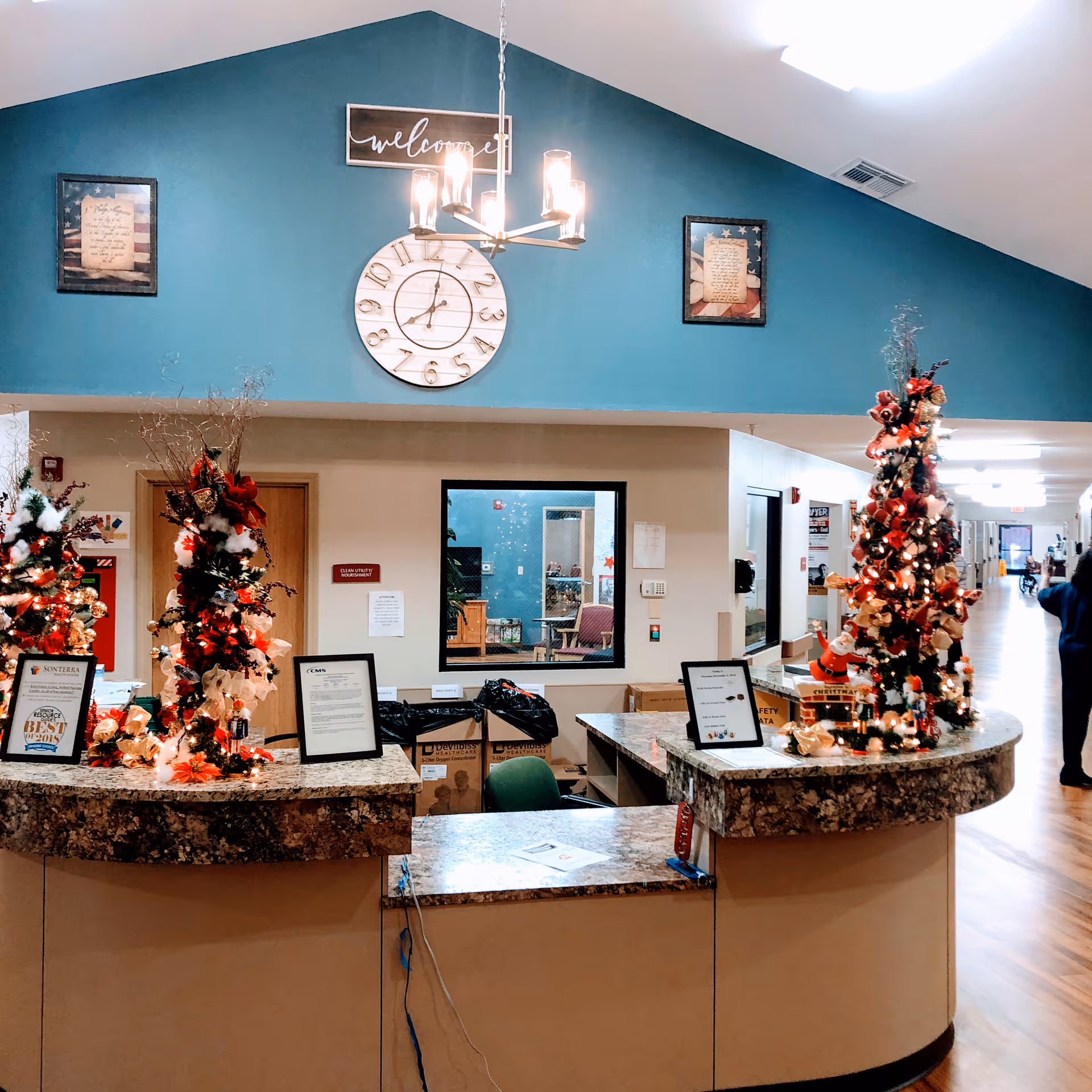 Reception desk area decorated with Christmas-themed decorations including small Christmas trees with lights and ornaments. Behind the desk is a blue wall with a large clock, a 'welcome' sign, and two framed pictures. The floor is wooden, and a hallway extends to the right with a person walking away in the distance.