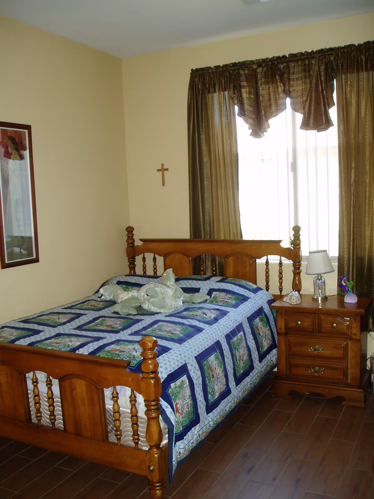 Sunlit bedroom with a wooden bed covered by a patterned quilt, a nightstand with a lamp, and a window with curtains.