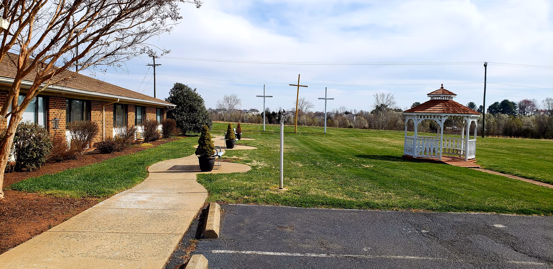 Outdoor view of a senior living facility with a brick building on the left, a paved walkway lined with potted plants, three crosses on poles in the grassy area, and a white gazebo with a red roof on the right under a partly cloudy sky.