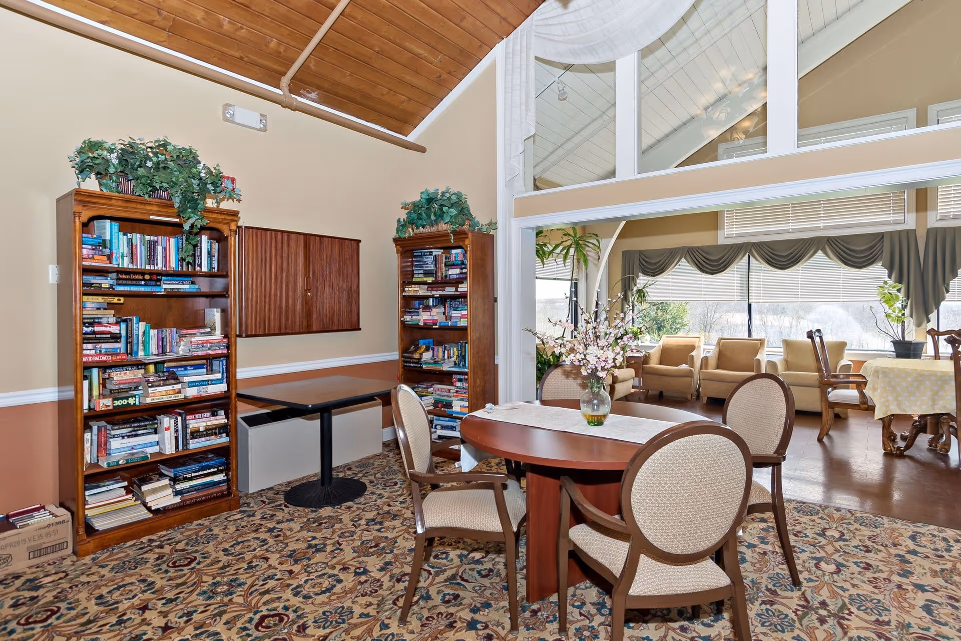 A cozy interior room with two wooden bookshelves filled with books, a round wooden table with four upholstered chairs, and a vase with flowers on the table. The room has a patterned carpet and a high wooden ceiling. In the background, there is a sunlit sitting area with armchairs and large windows with blinds and drapes.