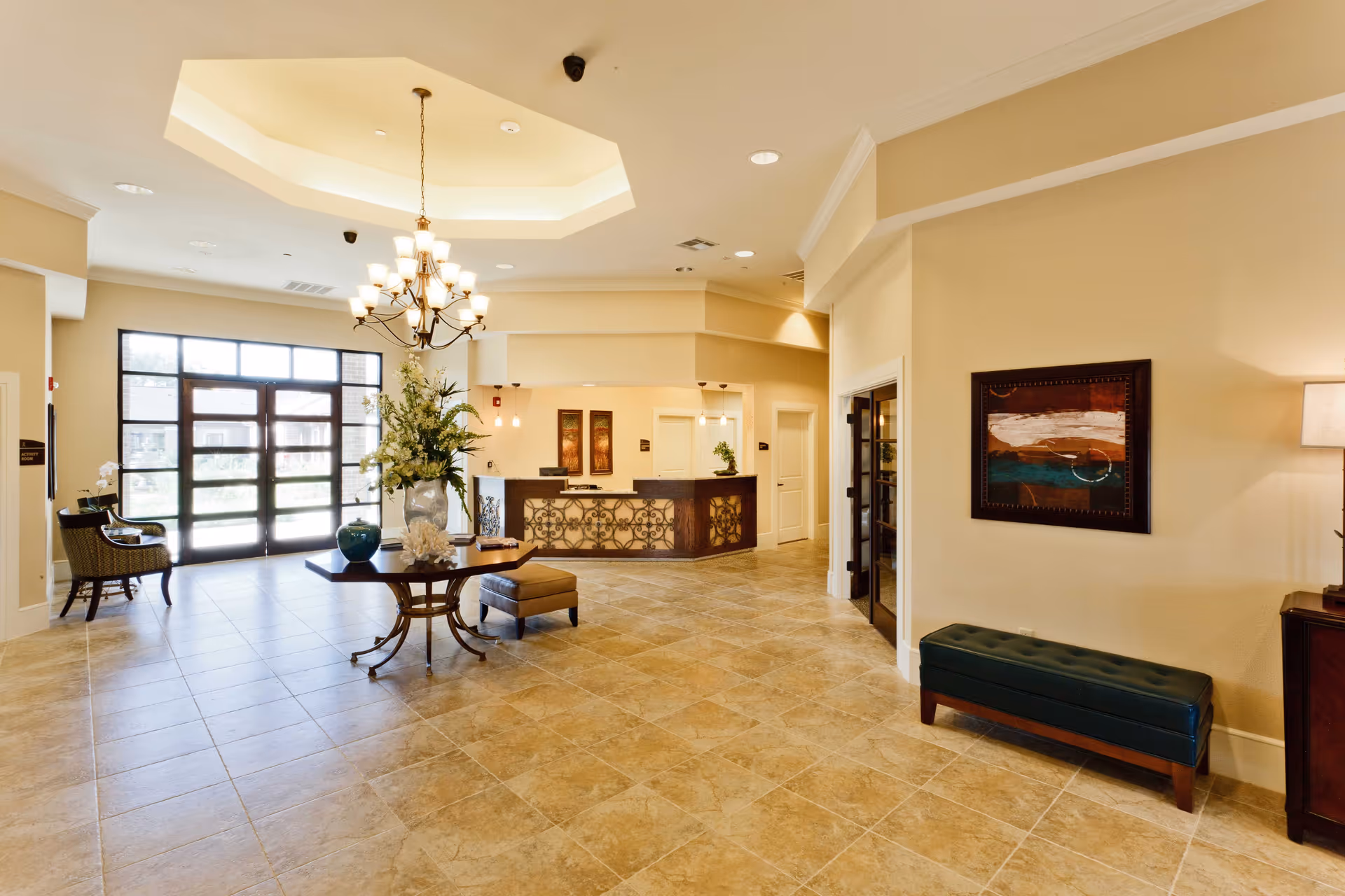 Spacious and well-lit assisted living facility lobby with tiled floors, a central table adorned with a large floral arrangement, a chandelier hanging from a recessed ceiling, a reception desk in the background, seating areas, and decorative artwork on the walls.