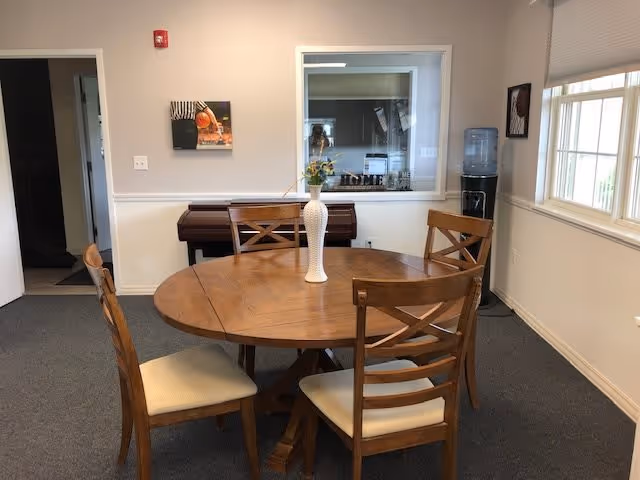 A small dining area with a round wooden table and four wooden chairs with cushioned seats. A white vase with flowers is placed on the table. In the background, there is a piano against the wall, a water cooler near a window, and a framed picture on the wall. The room has beige walls and carpeted flooring.