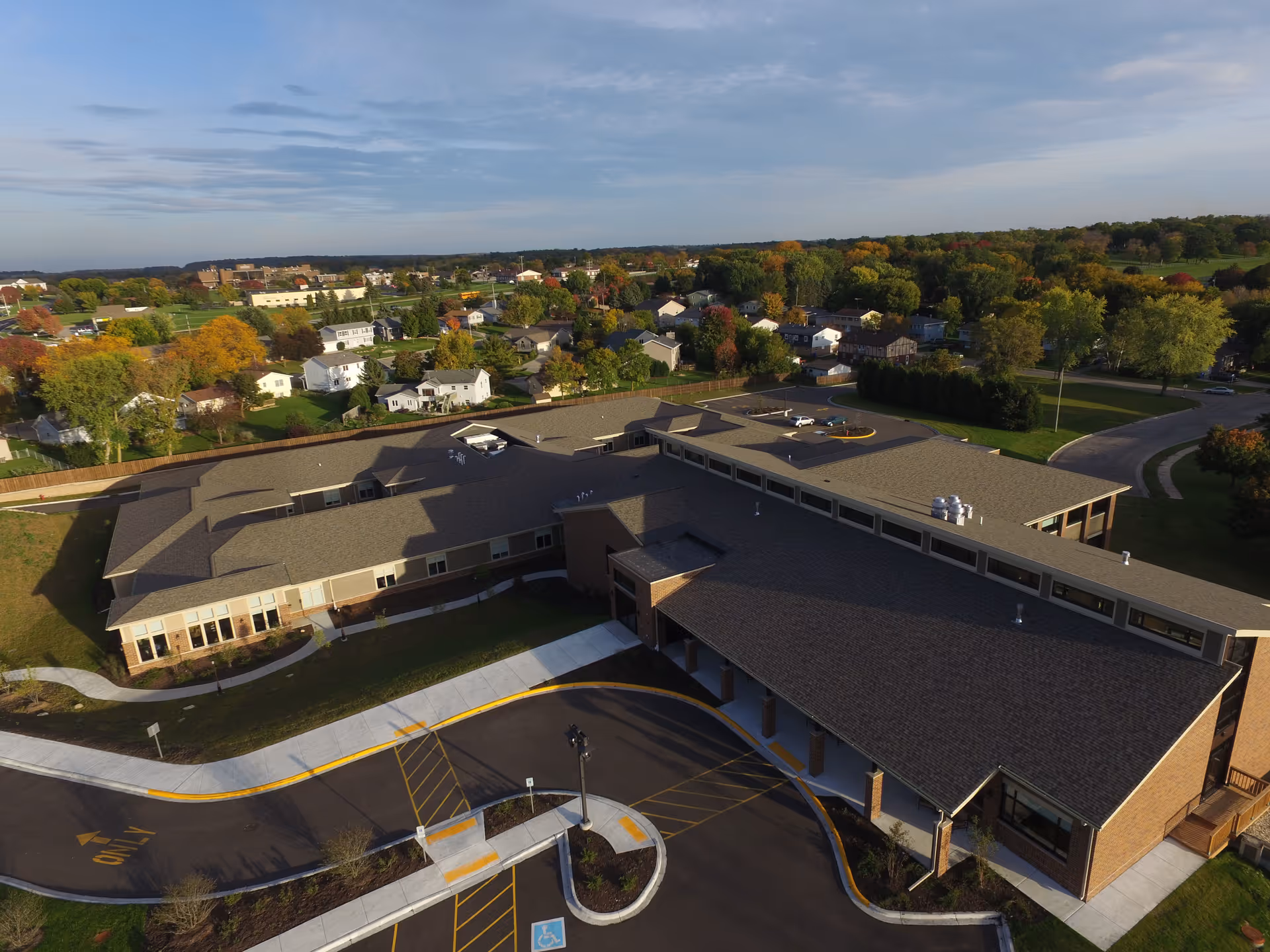 Aerial view of The Marquardt senior living facility showing a large single-story building with a brown roof surrounded by a curved driveway and parking area. The facility is located in a suburban area with houses, trees, and greenery visible in the background under a partly cloudy sky.
