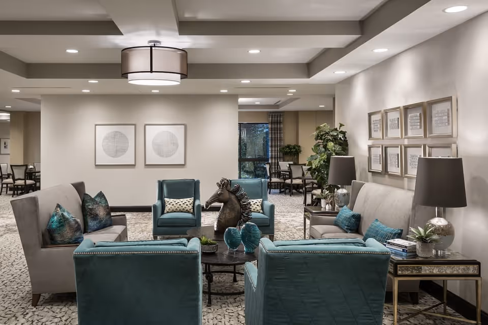 A well-lit living room area in a senior living facility with two gray sofas and four teal armchairs arranged around a round coffee table. The coffee table features decorative items including a horse head sculpture and blue vases. The walls are adorned with framed artwork, and there are two table lamps on side tables next to the sofas. In the background, there is a dining area with tables and chairs near a window with curtains.
