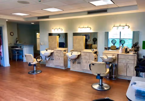 Interior view of a salon area in a senior living facility with three salon chairs in front of sinks and mirrors, wooden flooring, and a reception desk in the background.