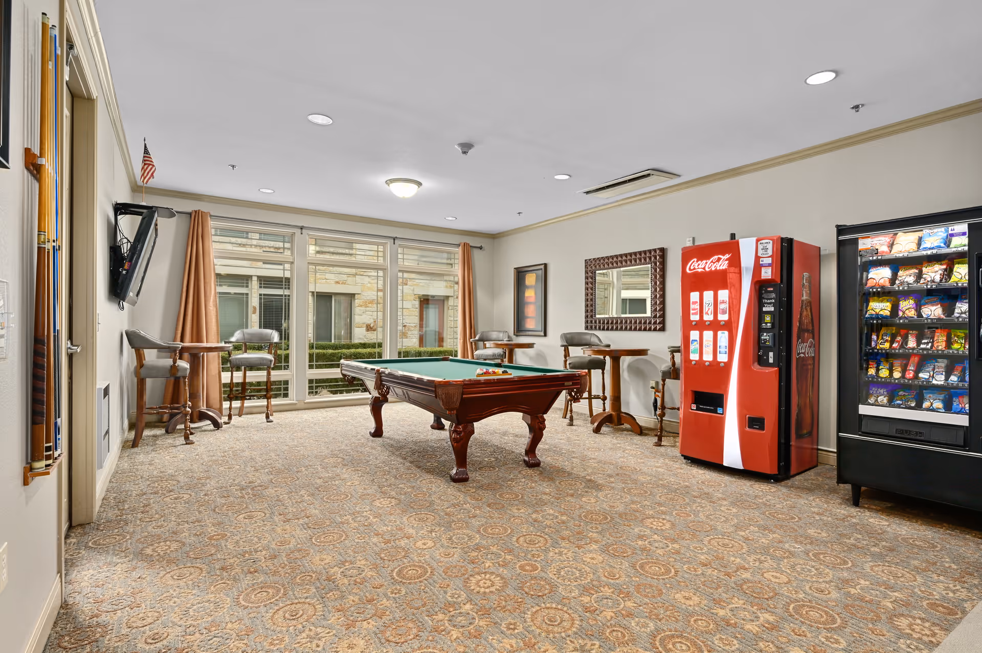 A recreational room with a pool table in the center, surrounded by tall chairs and small tables near large windows with beige curtains. On the right side, there is a Coca-Cola vending machine and a snack vending machine. The room has patterned carpet flooring and neutral-colored walls with a mirror and framed artwork.