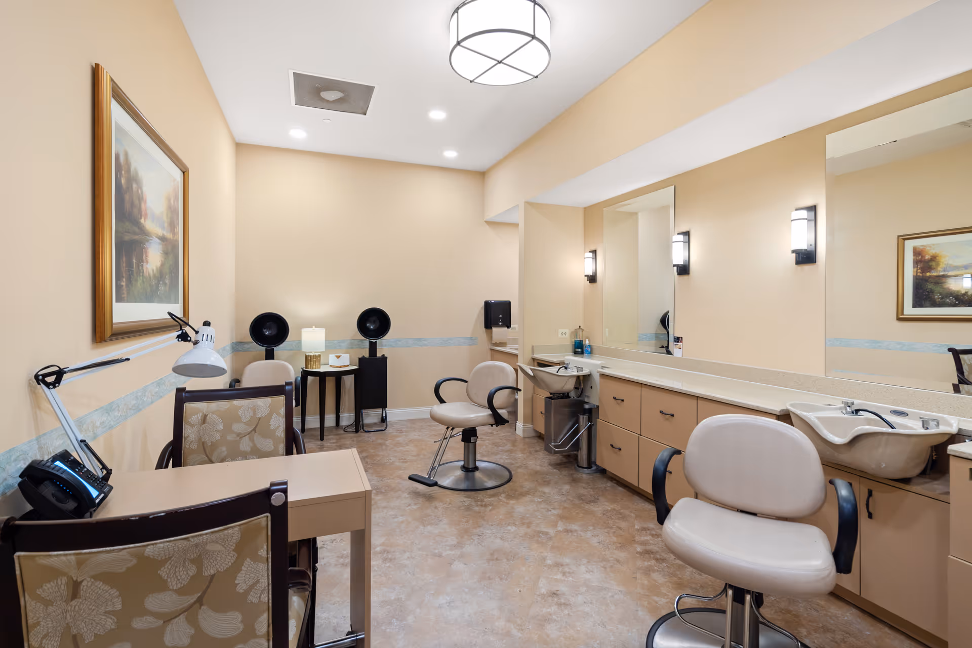 Interior view of a salon area in a senior living facility with beige walls and flooring. The room features two salon chairs in front of a long counter with sinks, mirrors, and cabinets. There is a small table with two black hair dryers and a lamp, as well as a desk with a telephone and a lamp. A framed landscape painting hangs on the wall.