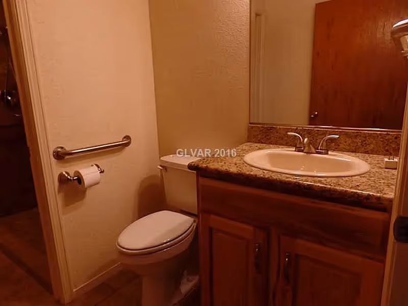 A small bathroom with a toilet, a wall-mounted grab bar, a toilet paper holder, a wooden vanity with a granite countertop, a sink with a faucet, and a large mirror above the sink. The bathroom door is partially visible in the background.