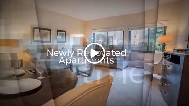 A newly renovated apartment living room with beige walls, a large window letting in natural light, a wooden dining table with chairs, a comfortable armchair, a lamp on a side table, and framed artwork on the wall.