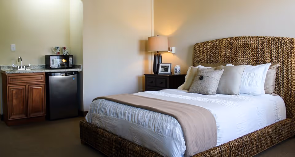 A cozy senior living bedroom featuring a large woven headboard bed with white and beige bedding and multiple pillows. To the left, there is a small kitchenette area with a sink, microwave, and mini refrigerator. A bedside table with a lamp, framed photo, and decorative item is positioned next to the bed.