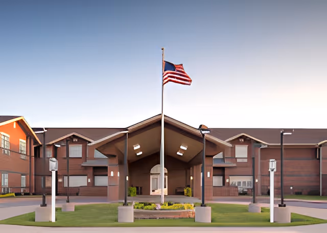 Front exterior view of Canoe Brook Assisted Living - Catoosa building with a large covered entrance, an American flag on a flagpole in the center, and well-maintained landscaping.