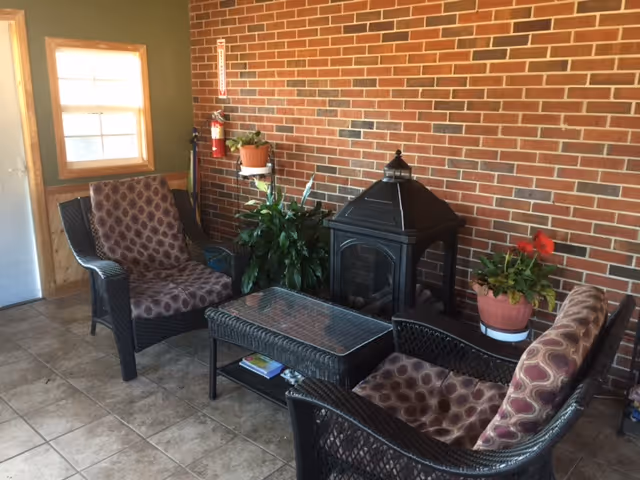 A cozy indoor seating area with two cushioned wicker chairs and a wicker loveseat arranged around a glass-top coffee table. Behind the seating is a black wood stove against a red brick wall. There are potted plants on either side of the stove, and a window with wooden trim is visible on the left side of the image.