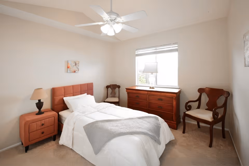A neatly made single bed with white bedding and a gray throw blanket in a bright bedroom. The room features a coral-colored headboard and matching nightstand with a lamp, two wooden chairs with cushions, a wooden dresser with a lamp on top, and a window with blinds letting in natural light.