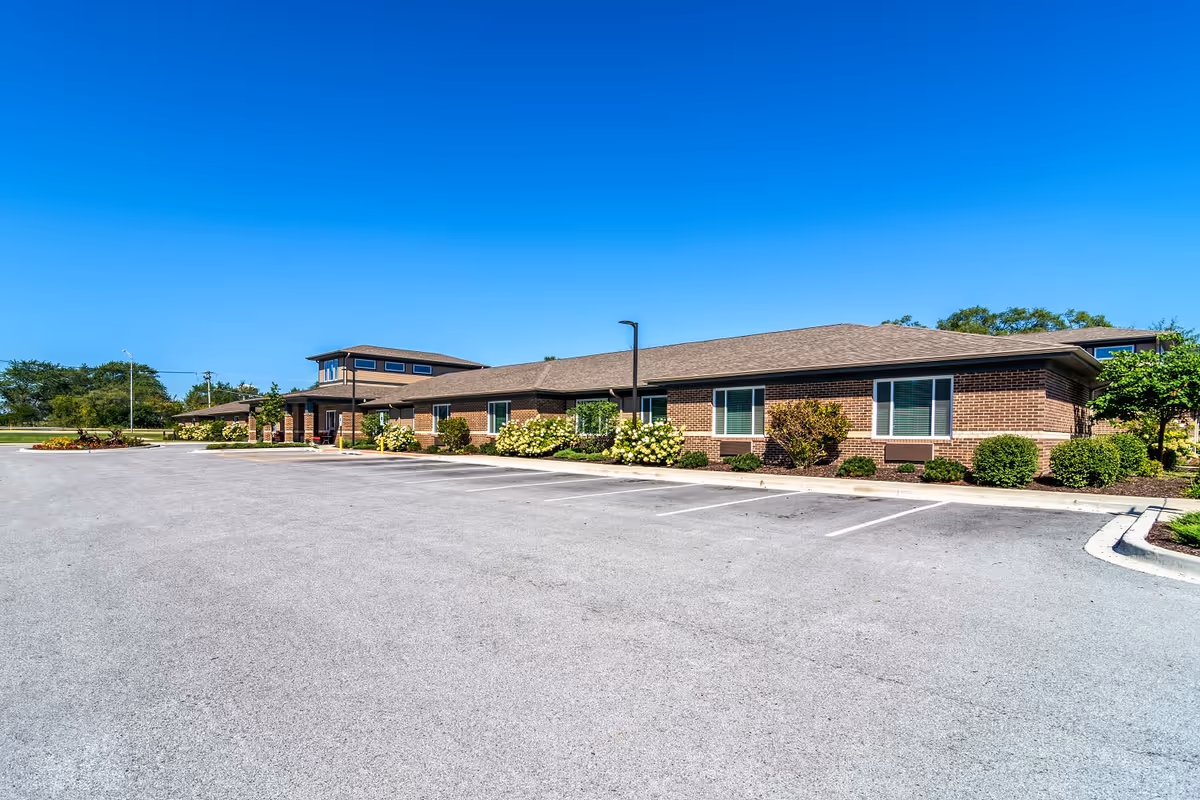 Exterior view of a single-story brick building with a large empty parking lot in front, clear blue sky, and some greenery and bushes around the building.