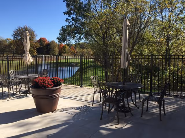 Outdoor patio with metal tables, chairs, closed umbrellas and a large potted flower overlooking a fenced pond and trees.