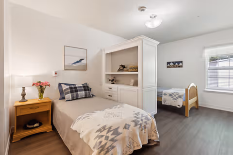 A bright and clean bedroom with two single beds separated by a white cabinet. Each bed has a patterned blanket and pillows. There is a wooden nightstand with a lamp and a vase of flowers next to one bed. The room has light-colored walls, a window with blinds, and framed artwork on the walls.