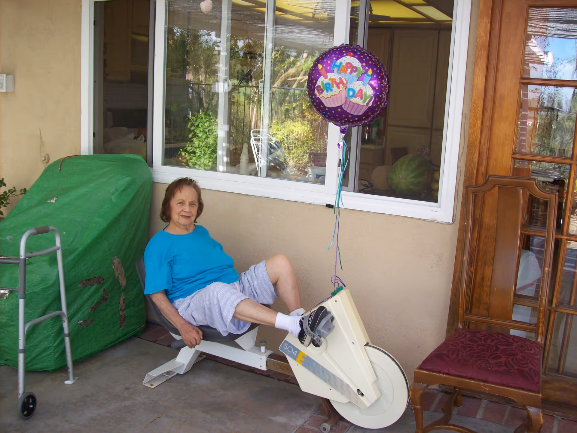 An elderly woman in a blue shirt and light shorts is sitting on a stationary exercise bike on a covered patio. A purple 'Happy Birthday' balloon is tied to the bike. There is a green covered object and a walker to the left, and a wooden chair with a red cushion to the right. A window and a wooden door are visible in the background.