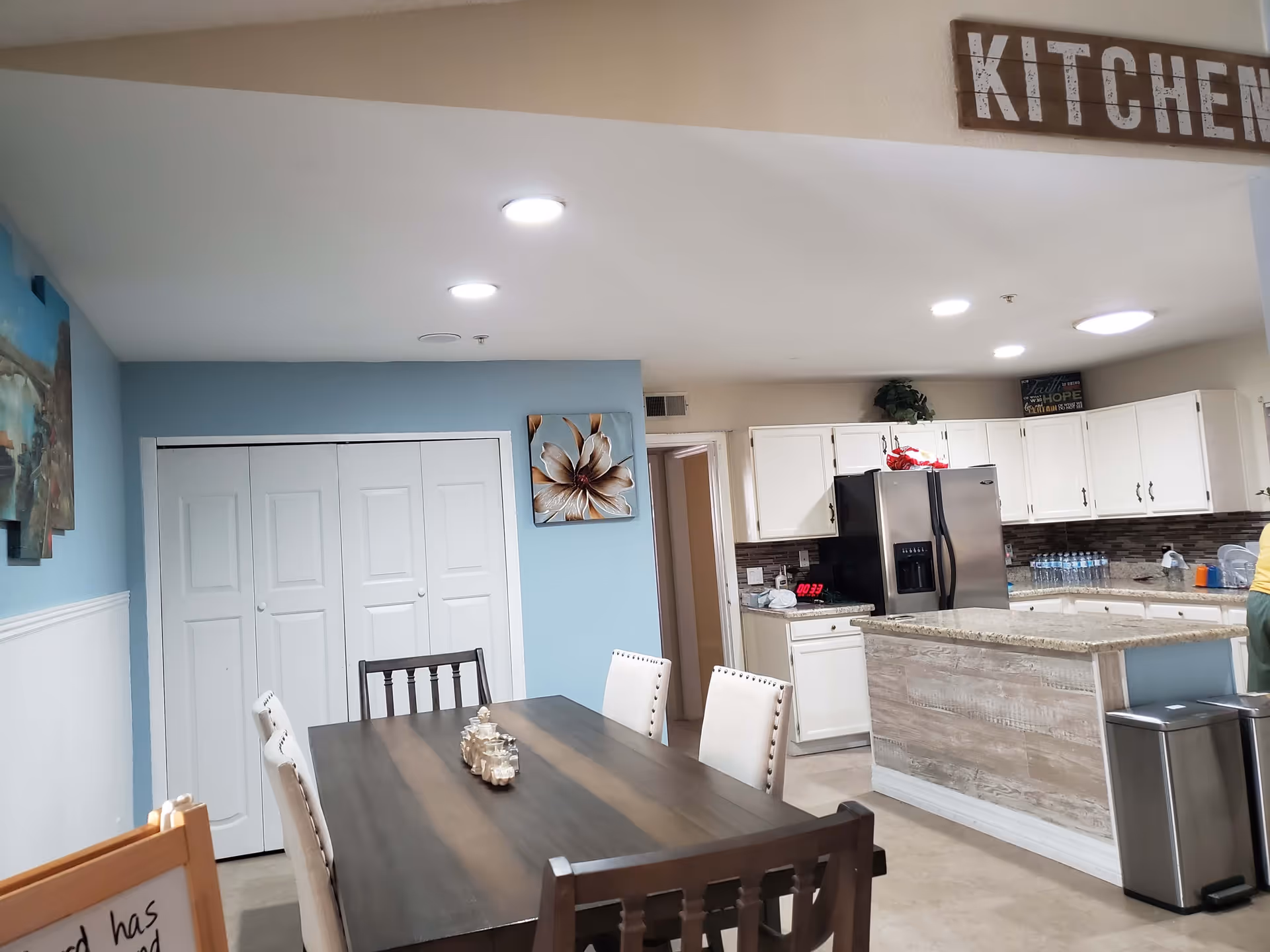 Interior view of a kitchen and dining area in a senior living facility. The dining area features a dark wooden table with six chairs, some upholstered in white fabric. The kitchen has white cabinets, a granite countertop island, a stainless steel refrigerator, and various kitchen items on the counters. The walls are painted light blue and beige, with decorative artwork and a wooden sign reading 'KITCHEN' above the kitchen entrance.
