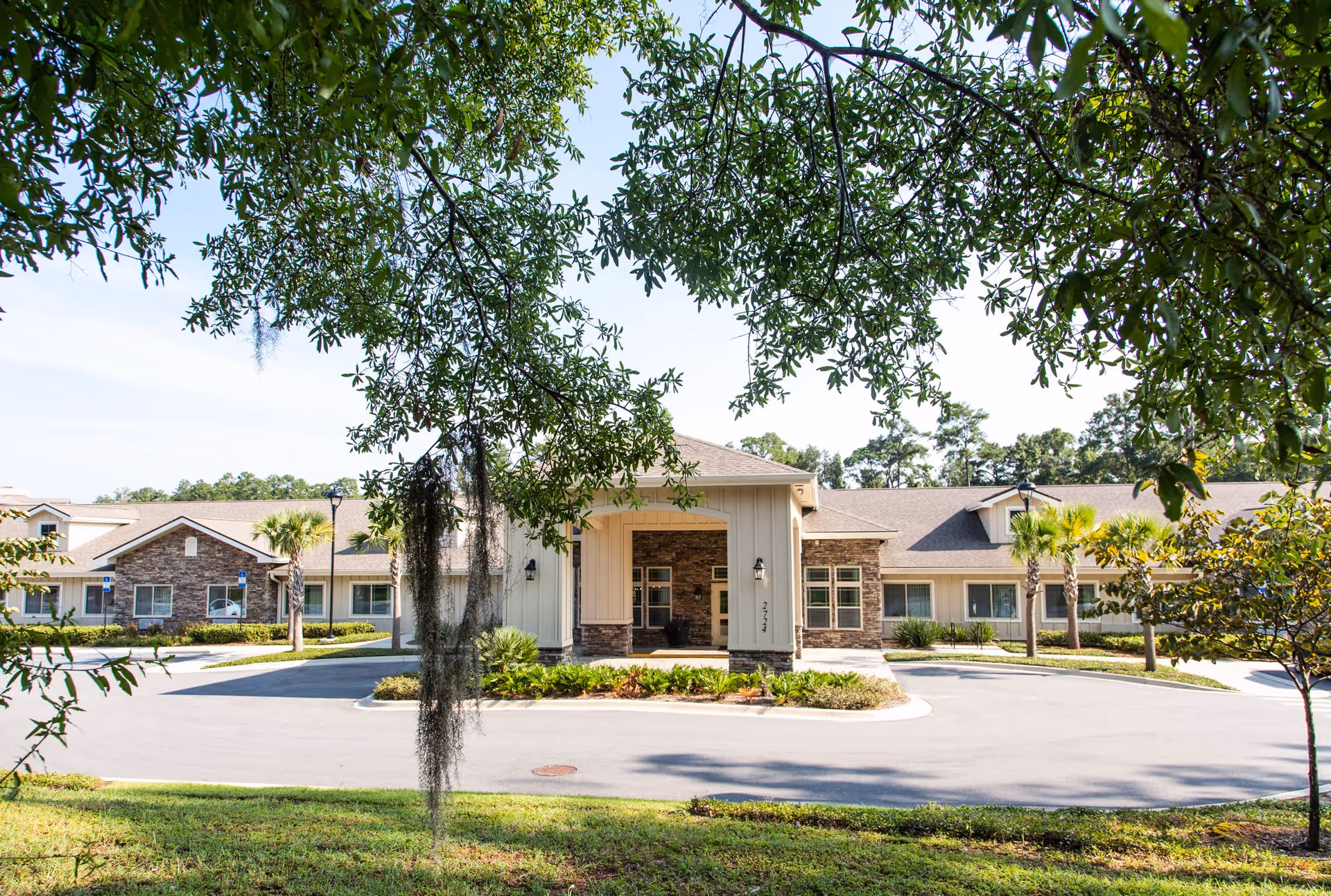 Front exterior view of Azalea Gardens Assisted Living & Memory Care building with a circular driveway, landscaped greenery, palm trees, and large tree branches framing the top of the image.