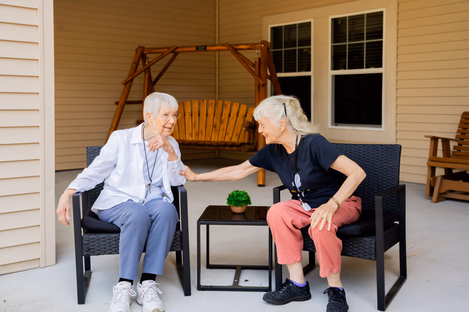 Two elderly women sitting on black wicker chairs on a covered patio, smiling and interacting with each other. There is a small black table with a small potted plant between them, a wooden swing in the background, and beige siding with two windows behind them.