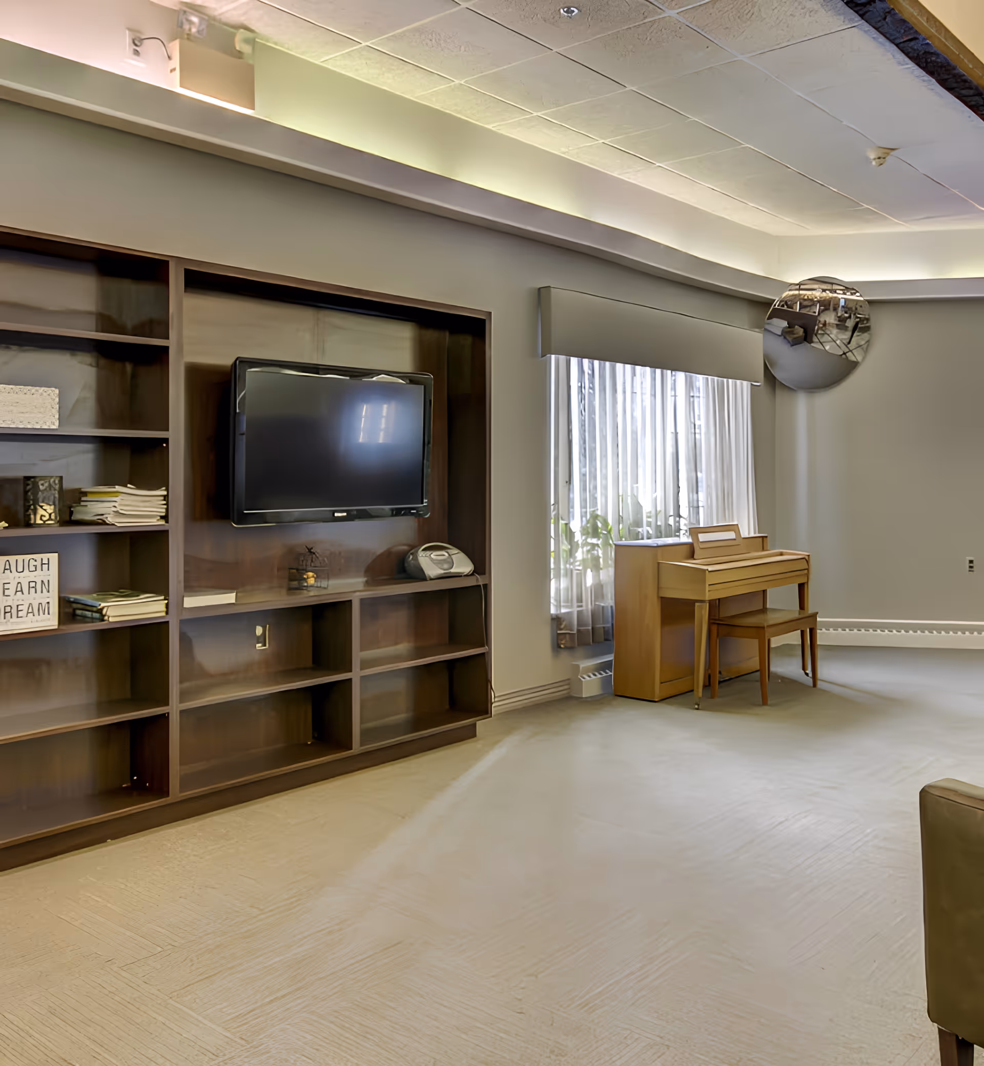 A living room area in a senior living facility featuring a wall-mounted flat-screen TV set within a wooden shelving unit, a small wooden piano with a matching bench near a window with sheer curtains, and a convex mirror mounted on the wall reflecting part of the room.