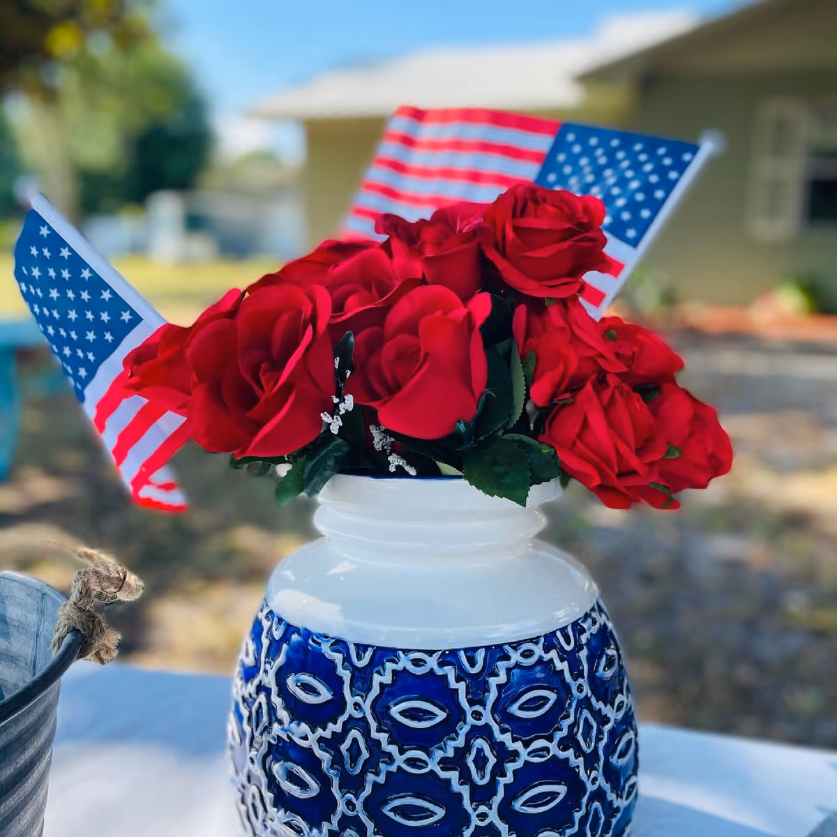 A decorative blue and white vase filled with red roses and two small American flags, placed on a white surface outdoors with a blurred background of greenery and a building.