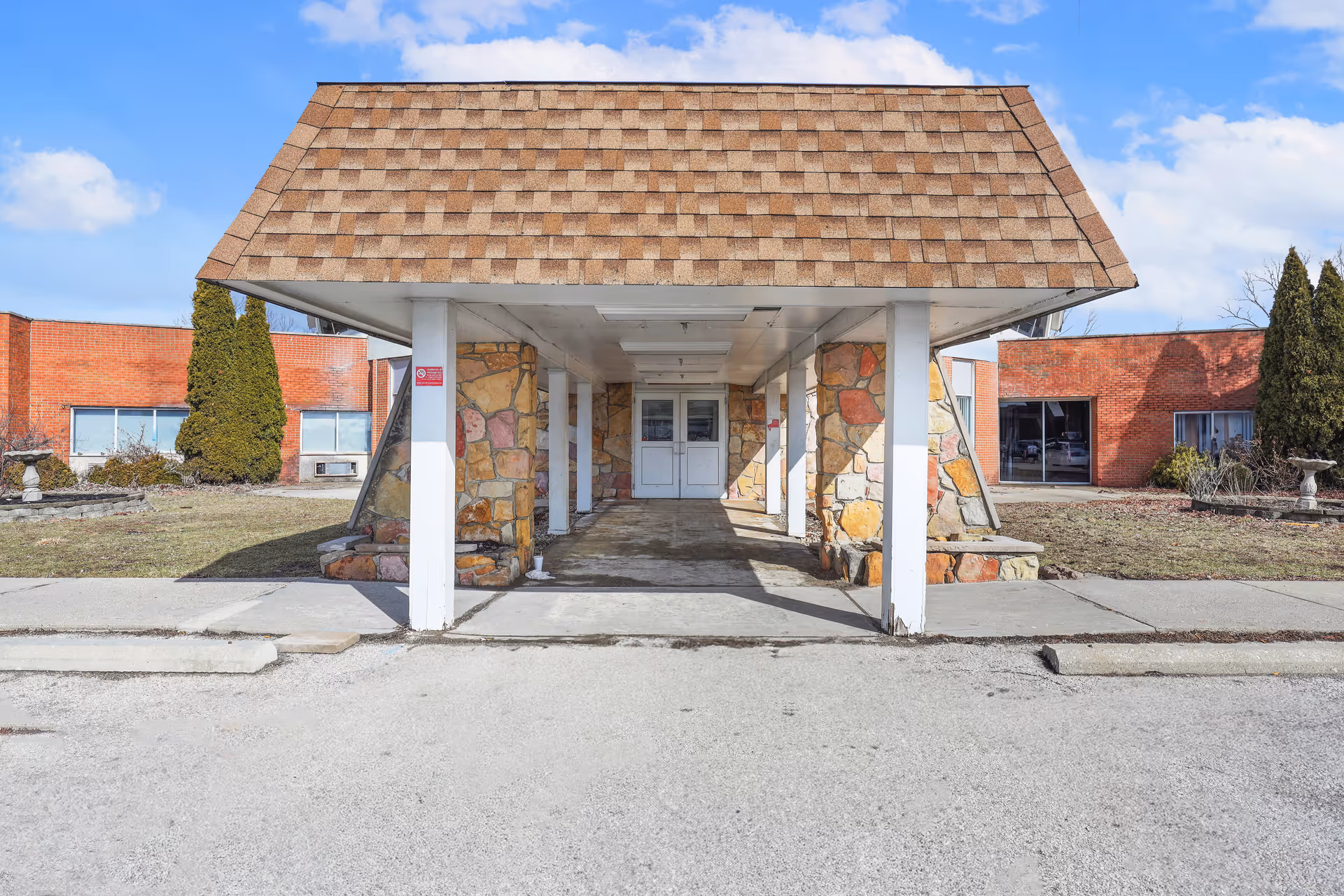 Entrance of a healthcare facility with a covered driveway supported by white pillars and stone walls. The building has a shingled roof over the entrance and red brick walls on either side. There are some shrubs and small trees in the surrounding grassy area under a partly cloudy blue sky.