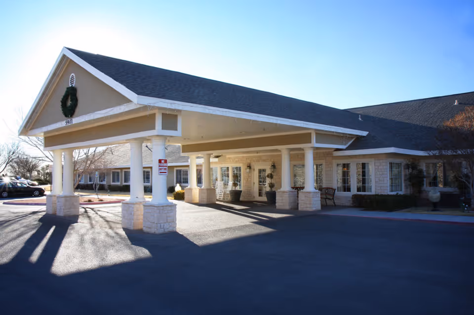 Front entrance of a single-story senior living facility with a covered porte-cochère, white columns, stone facade and driveway under a clear sky.