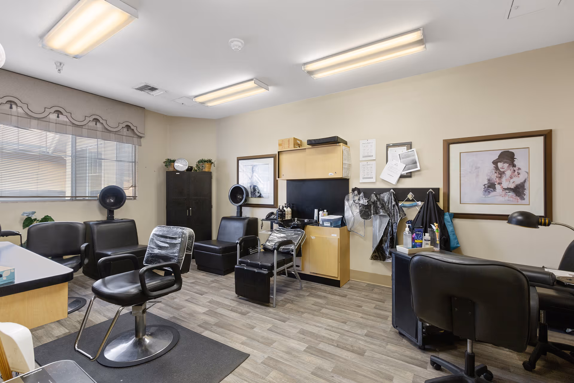 Interior view of a hair salon area within a senior living facility. The room features several black salon chairs, hair drying stations, a cabinet with hair care products, and framed artwork on the walls. The floor is wood-patterned, and the room is lit by fluorescent ceiling lights.