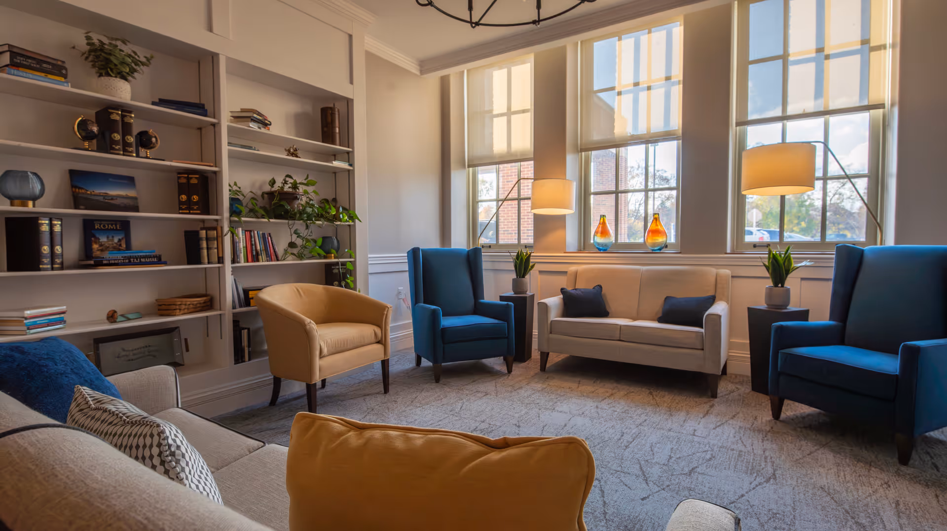 A bright and cozy living room with large windows letting in natural light. The room features a beige sofa with dark blue pillows, two blue armchairs, and a tan armchair arranged around a carpeted floor. There are two floor lamps with beige shades, potted plants on small tables, and a built-in bookshelf filled with books and decorative items.
