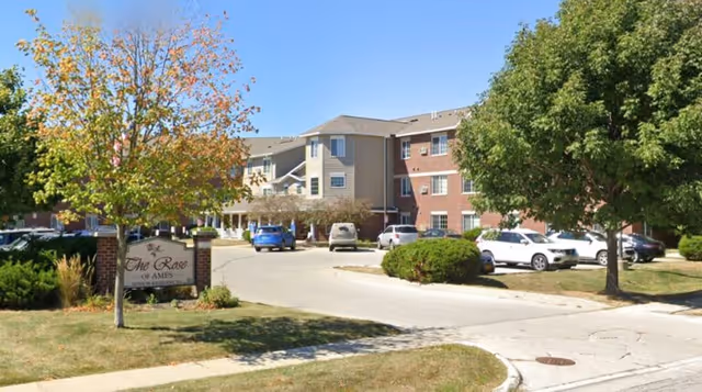 Exterior view of The Rose of Ames senior living facility showing a multi-story building with a mix of brick and siding, surrounded by trees and a parking area with several cars.