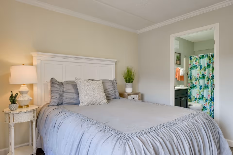 A bedroom with a neatly made bed featuring gray bedding and pillows. To the left of the bed is a small round white side table with a lamp and a small plant. To the right of the bed is a wooden nightstand with a potted plant. An open door reveals a bathroom with a colorful tropical shower curtain, a toilet, and a vanity with a sink and towels.