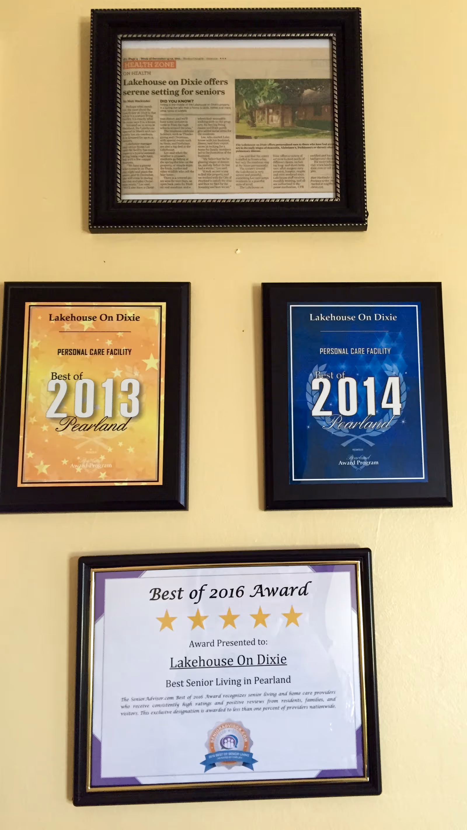 Four framed awards and a newspaper article displayed on a beige wall. The awards recognize Lakehouse On Dixie as the best personal care facility in Pearland for the years 2013, 2014, and 2016. The newspaper article highlights Lakehouse On Dixie offering a serene setting for seniors.