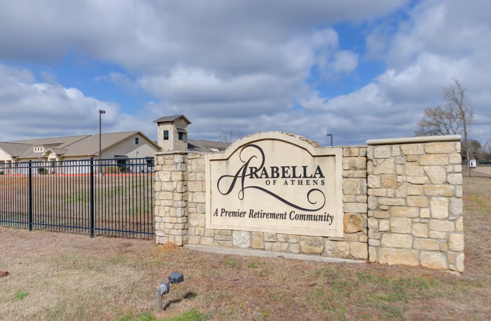 Stone sign at the entrance of Arabella of Athens Senior Living, a premier retirement community, with a building and black metal fence in the background under a partly cloudy sky.