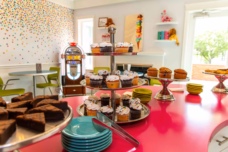 A colorful dining area with a pink countertop displaying a variety of desserts including cupcakes with white frosting and sprinkles, chocolate brownies, and muffins. There are stacks of turquoise and yellow plates, a vintage jukebox in the background, green chairs around tables, and a wall decorated with multicolored dots and artwork.