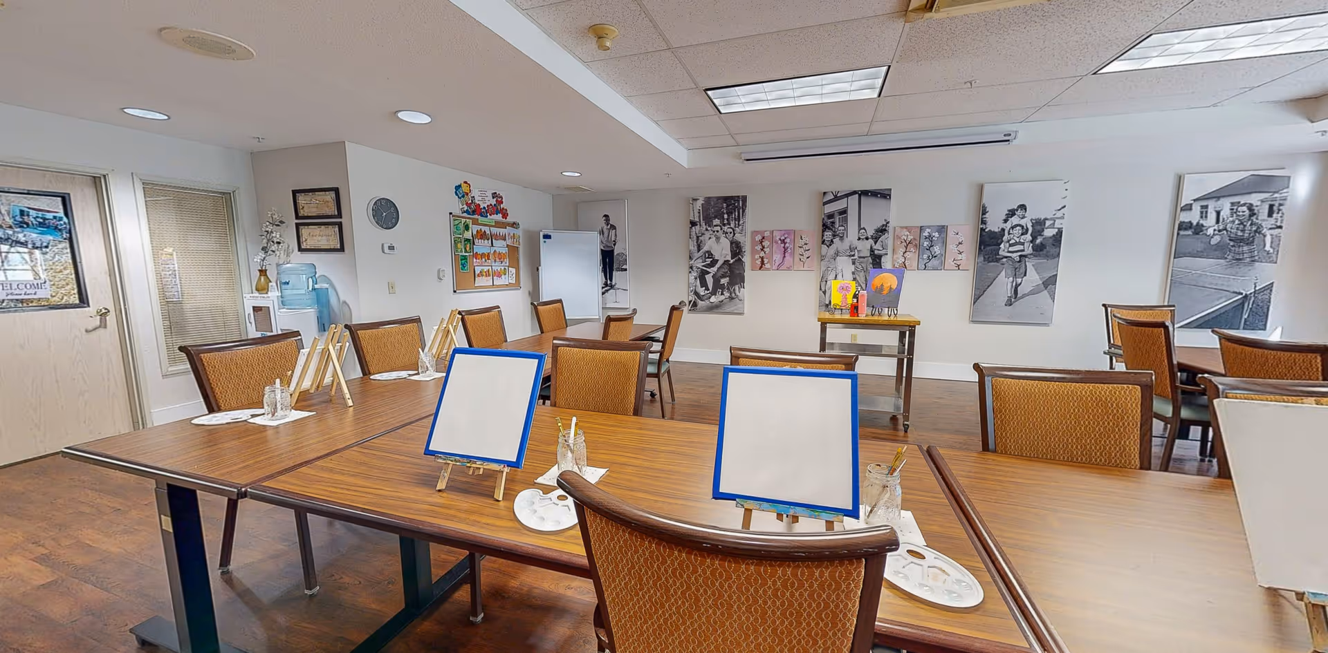 A well-lit activity room with several tables and chairs arranged for painting. Each table has small easels with blank canvases, paint palettes, and jars with brushes. The walls are decorated with black and white photos and colorful artwork. There is a water cooler and a bulletin board on one side of the room.