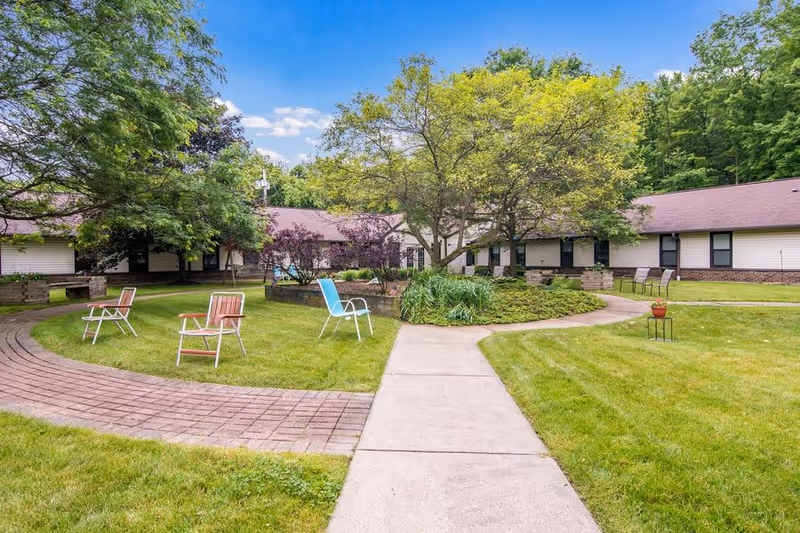 Outdoor courtyard area at Celebration Villa of Allison Park with a paved walkway, green grass, several trees, and a few chairs placed around the lawn. The building surrounds the courtyard with white siding and brown roofs under a blue sky with some clouds.