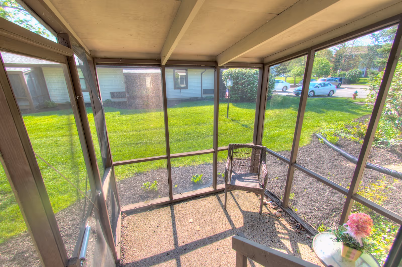 A small screened-in porch area with a single wicker chair and a round table holding a potted pink flower. The porch overlooks a grassy lawn with bushes and a parking lot with several cars in the background.