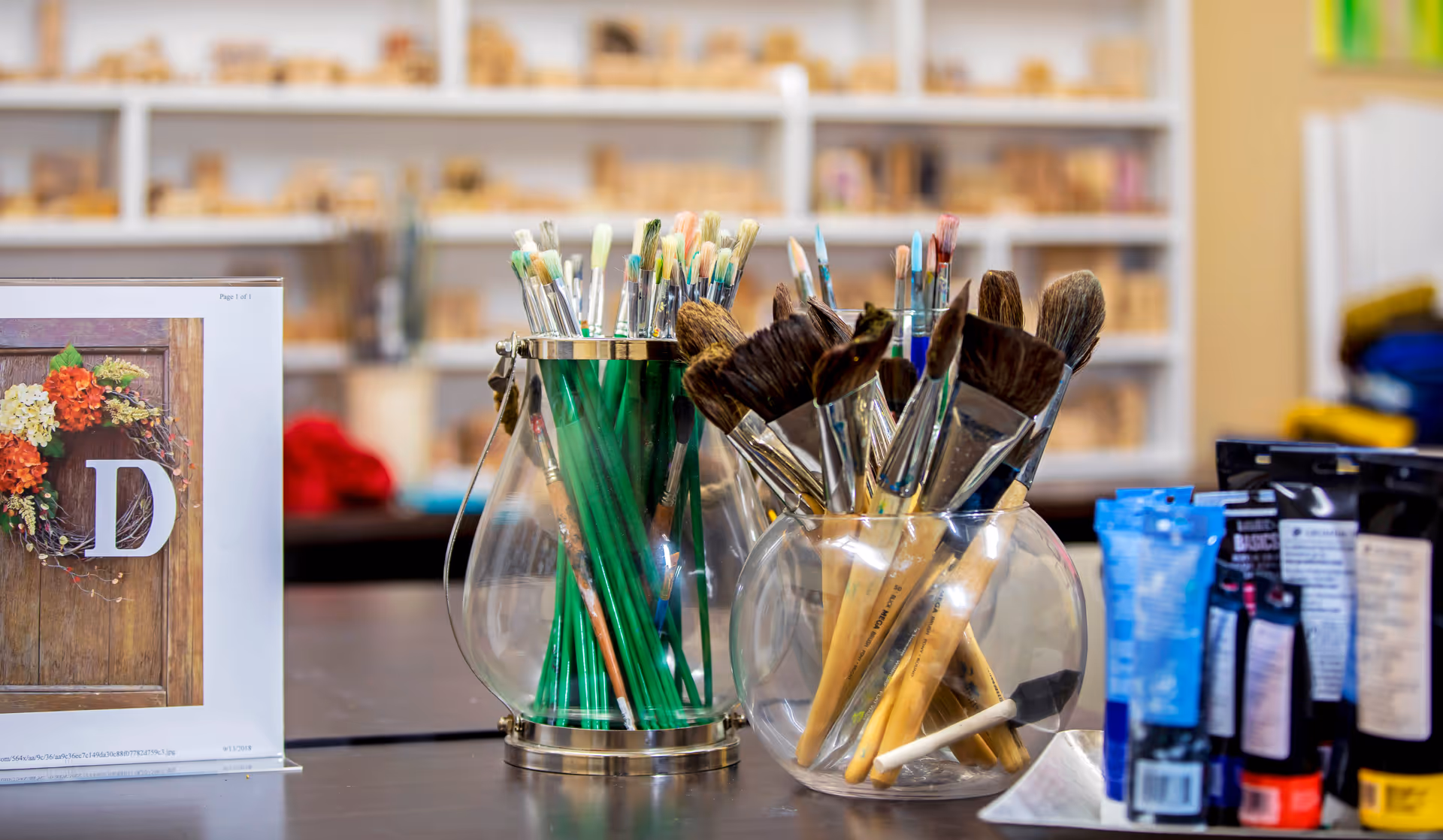 Two glass containers filled with various paintbrushes on a table, with paint tubes and a printed image of a wooden door with a floral wreath and the letter D on the left side. Shelves with wooden blocks or craft materials are visible in the blurred background.