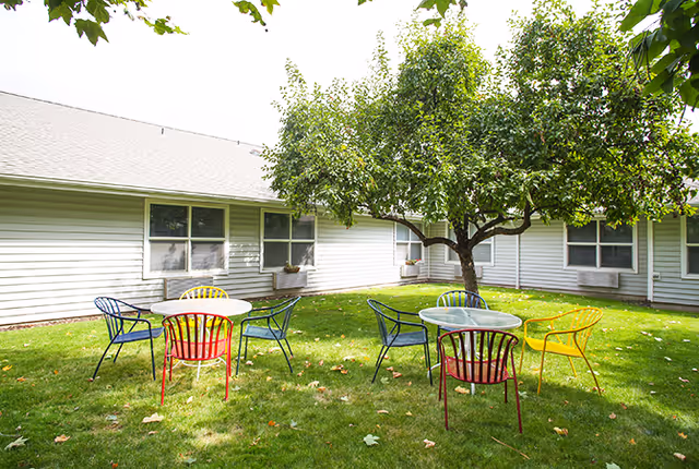Sunny courtyard with round tables and colorful metal chairs arranged on grass beneath a tree beside a single-story building.