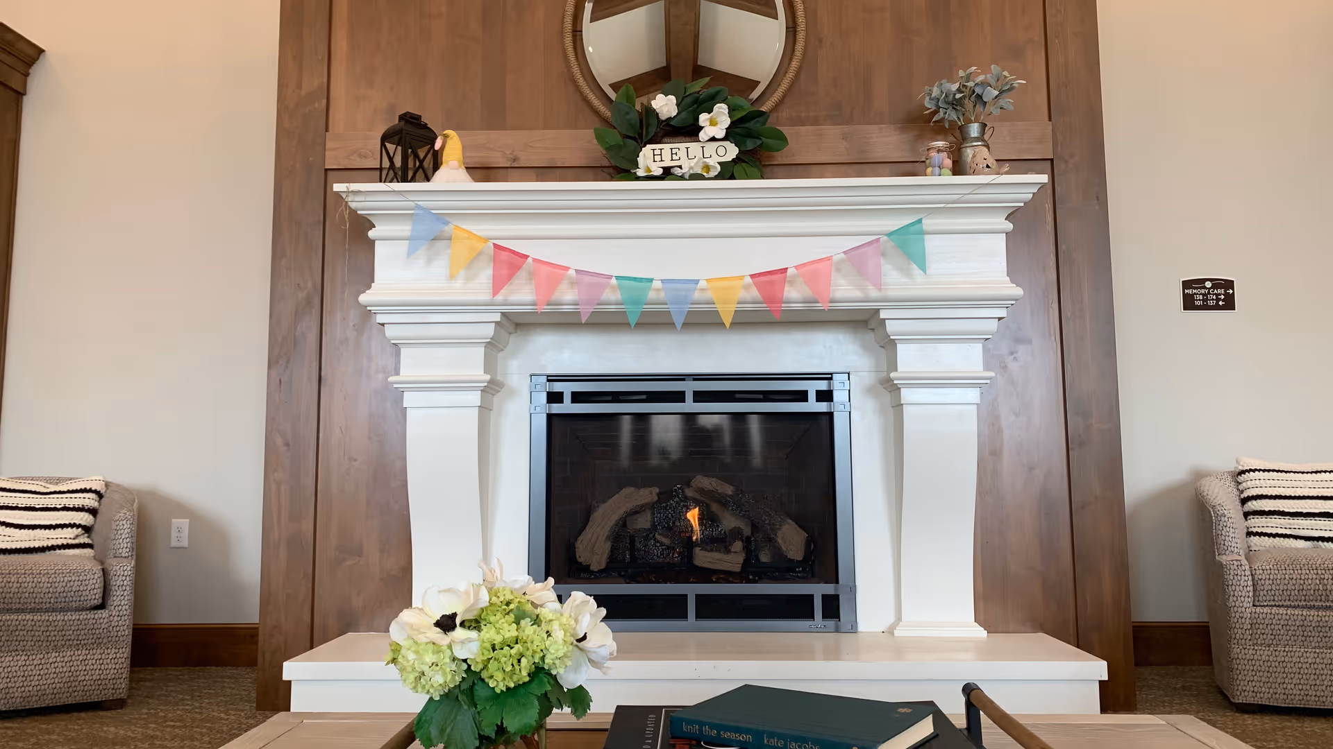 A cozy living room area featuring a white fireplace with a small fire burning inside. The mantel is decorated with a round mirror, a 'HELLO' sign surrounded by greenery and flowers, a small lantern, a duck figurine, and two small jars with flowers. A colorful triangular pennant banner hangs across the mantel. In front of the fireplace is a white surface with a bouquet of flowers and an open book. Two cushioned armchairs with striped pillows are partially visible on either side of the fireplace.