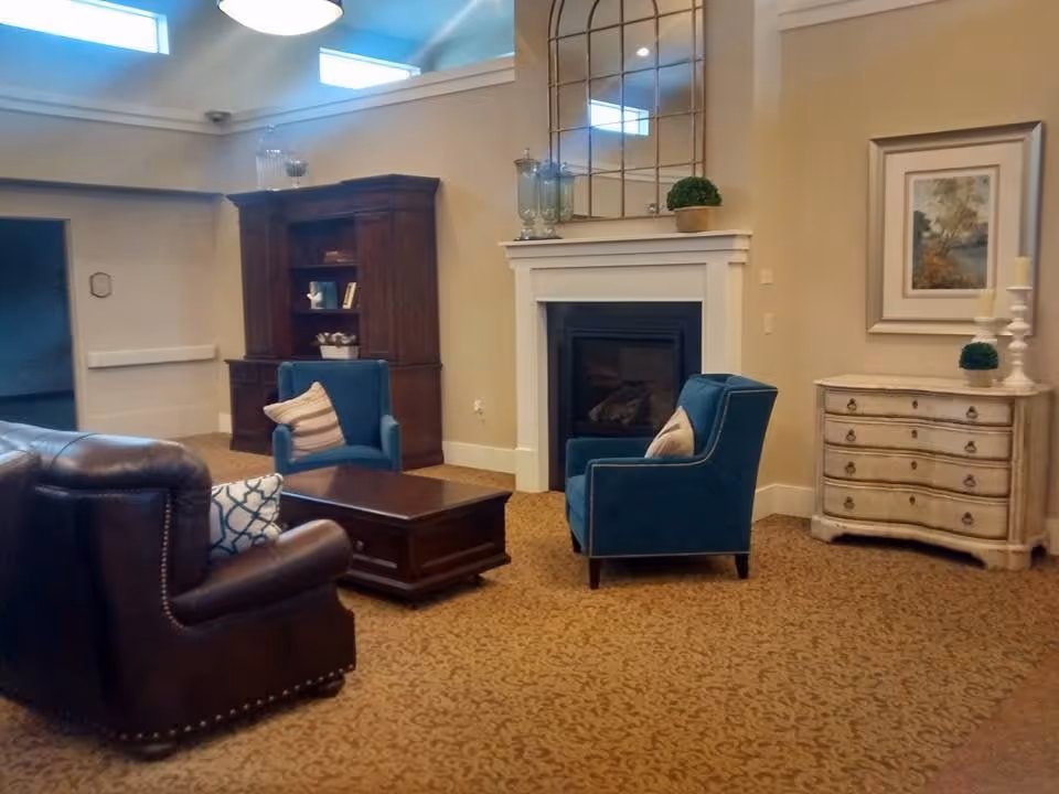 A cozy living room area with a brown leather armchair, two blue upholstered armchairs with pillows, a wooden coffee table, a white fireplace with decorative items on the mantle, a large wooden cabinet, and a small white chest of drawers with a framed picture and candles on top. The room has beige walls and patterned carpet flooring.
