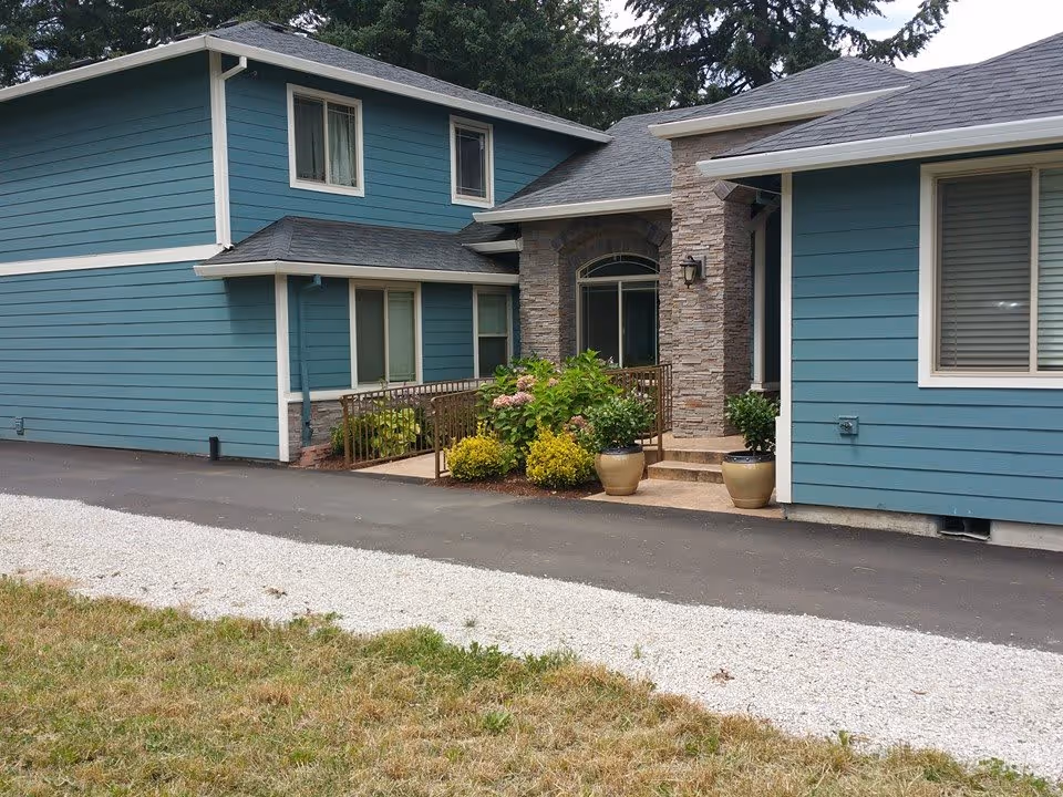 Exterior view of a blue two-story building with white trim and a stone entrance. There is a paved driveway and a gravel path in front of the building, along with some green shrubs and potted plants near the entrance.