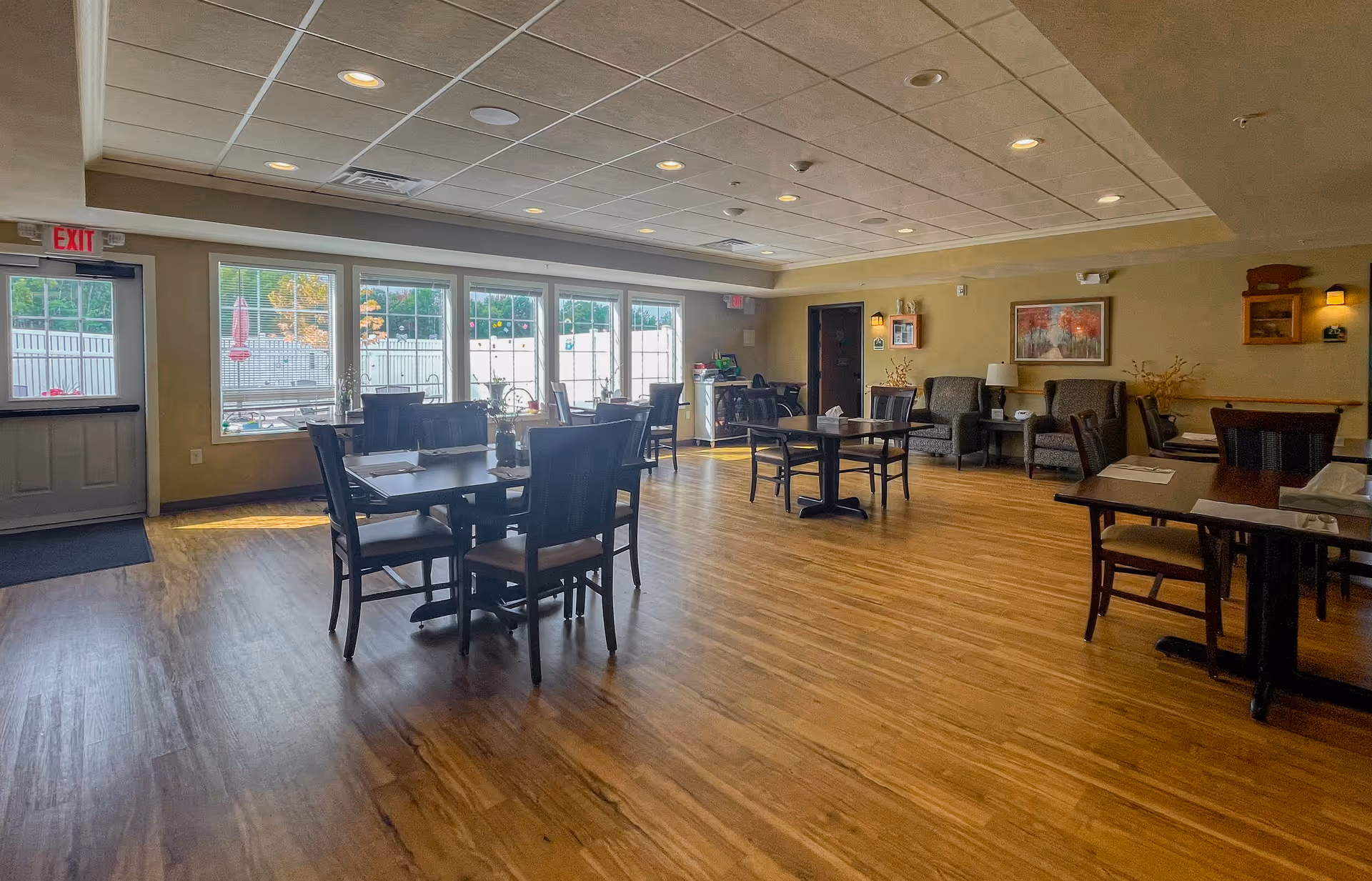 A spacious dining room with wooden floors, several tables and chairs arranged neatly, large windows letting in natural light, and a seating area with armchairs and a side table against the far wall.